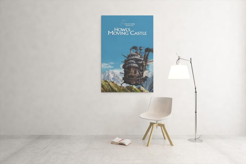 May include: A poster of Howl's Moving Castle hangs on a white wall. The poster features the animated castle against a blue sky and mountains. A white chair, a book, and a lamp are in front of the wall.