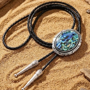 May include: A bolo tie featuring a braided black leather cord and a central oval-shaped pendant. The pendant has an iridescent abalone shell inlay set in a silver-toned metal frame with etched details. Two silver-toned metal tips finish the cord.