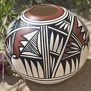May include: A round, hand-painted ceramic pot with a wide opening. The pot features a cream-colored base with geometric designs in black, brown, and white. The rim is brown, and the overall design is symmetrical and detailed.