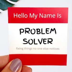 May include: A square adhesive name tag with a red border at the top and bottom. The white centre features the text Hello My Name Is in red, followed by the words PROBLEM SOLVER in bold black letters and fixing things no one else notices below.