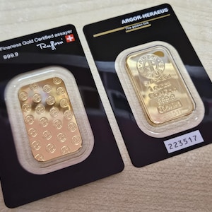May include: Two gold bars, each encased in a black and white card. One bar is stamped with "1 oz 999.9" and the other with "ARGOR-HERAEUS". The bars are rectangular with a repeating circular pattern.
