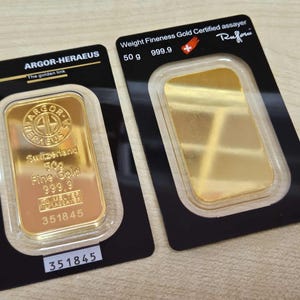 May include: Two gold bars, each in a black and clear plastic case. One bar is stamped with "ARGOR-HERAEUS" and "Switzerland 50g Fine Gold 999.9". The other bar is stamped with "50 g 999.9" and the word "Rufoni".