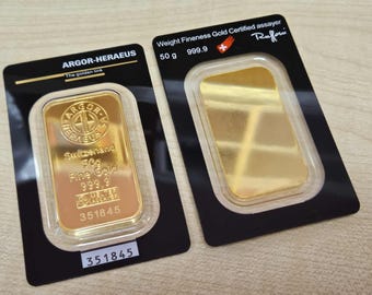 Puede incluir: Dos lingotes de oro, cada uno en una funda de plástico negro y transparente. Uno está estampado con "ARGOR-HERAEUS" y "Switzerland 50g Fine Gold 999.9". El otro está estampado con "50 g 999.9" y la palabra "Rufoni".