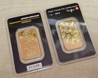 Puede incluir: Dos lingotes de oro, cada uno de 1 onza, presentados en envases de plástico transparente. Una barra está estampada con el texto "ARGOR-HERAEUS" y "Switzerland 1 ounce Fine Gold 999.9". La otra barra está estampada con "Ruffoni".