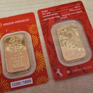 May include: Two gold bars in red packaging. One bar is stamped with the text "ARGOR-HERAEUS" and "1 ounce Fine Gold 999.9". The other bar is a limited edition "Year of the Horse 2026" with a horse design and the text "1 oz 999.9".