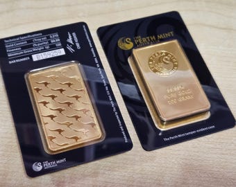 Puede incluir: Dos lingotes de oro de The Perth Mint, Australia, presentados en embalaje negro. Una barra presenta un patrón de canguros, la otra está estampada con el logotipo de la casa de la moneda y dice "99,99% Oro puro 100 GRAMOS". El embalaje incluye especificaciones técnicas.