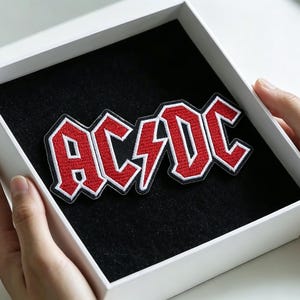 May include: An embroidered fabric patch featuring the red and white AC/DC logo with a central lightning bolt symbol. The patch is displayed inside a square white box with a black velvet-like interior lining, showcasing the detailed stitching.