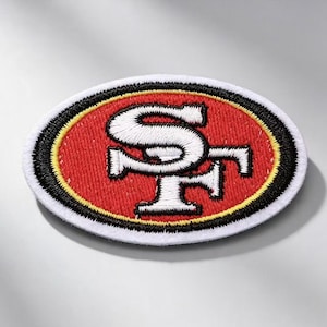 Puede incluir: Un parche bordado de forma ovalada con el logotipo de los San Francisco 49ers. El diseño incluye un monograma SF blanco entrelazado sobre un fondo rojo, rodeado por un borde amarillo fino y un borde negro grueso, con un acabado de borde blanco.