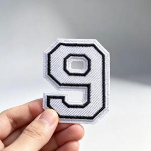 May include: A white embroidered patch in the shape of the number nine, featuring a bold black stitched border and a black inner outline. The patch has a textured, fabric-like surface with a clean, geometric design and a white stitched edge.
