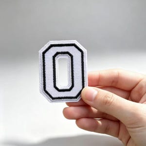 Può includere: Una toppa ricamata a forma di numero zero, caratterizzata da un centro bianco e un bordo nero spesso. La toppa presenta un aspetto testurizzato e cucito con un bordo esterno ottagonale netto e un'apertura rettangolare al centro.