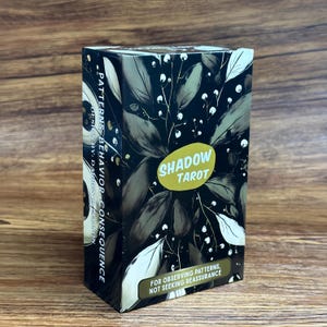 Puede incluir: Una caja rectangular de cartas de tarot con una ilustración botánica oscura de hojas y bayas blancas. El frente muestra un óvalo amarillo con el texto SHADOW TAROT y una etiqueta inferior que dice FOR OBSERVING PATTERNS, NOT SEEKING REASSURANCE.
