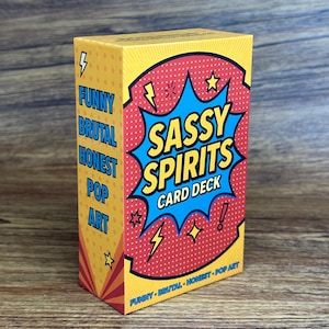 Könnte beinhalten: Eine rechteckige Kartenbox mit einer gelben Seitenfläche, die den Text FUNNY BRUTAL HONEST POP ART trägt. Die Vorderseite zeigt ein rotes Comic-Punktmuster mit einer blauen Sternform, die den Text SASSY SPIRITS CARD DECK enthält.