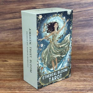 May include: A rectangular tarot deck box featuring the title Ethereal Bloom Tarot and the text Awaken. Trust. Bloom. Design by: Karol Robayo. The front displays an illustration of a figure in a flowing dress dancing before a large moon.