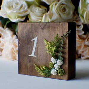 Può includere: Un segnaposto in legno con il numero 1 dipinto di bianco. Il blocco è decorato con verde artificiale e fiori bianchi e verdi. Il legno ha una finitura marrone naturale.