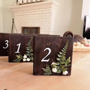Può includere: Segnaposto numerici da tavolo in legno marrone scuro, ciascuno con un numero bianco e decorazioni floreali. I numeri 1, 2 e 3 sono visibili. I segnaposto sono adornati con felci verdi e fiori bianchi, creando un'estetica rustica.