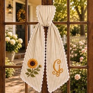 May include: A beige and white striped fabric door hanger featuring a white lace trim along the edges. One side displays an embroidered sunflower with green leaves, while the other side features an embroidered golden yellow letter G in a script font.