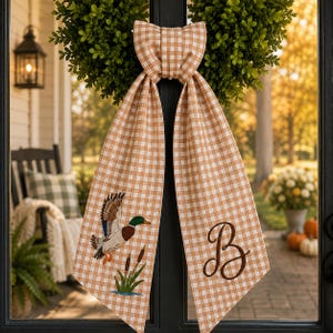 May include: A tan and white gingham fabric bow with two long hanging tails. One tail features an embroidered mallard duck in flight above green cattails, while the other tail displays a large, dark brown embroidered cursive letter B.