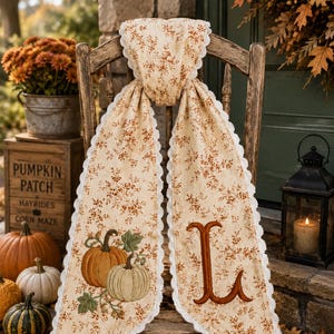 May include: A long, cream-colored fabric runner with a delicate brown floral pattern and white scalloped lace trim. One end features an embroidered orange and white pumpkin design with green leaves, while the other end displays a large, brown letter L.