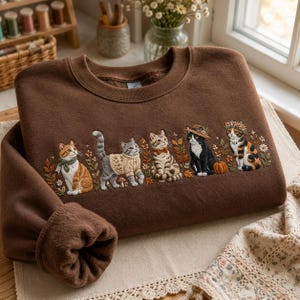 May include: A brown crewneck sweatshirt featuring detailed embroidery of five cats. The cats are depicted with various accessories including a green scarf, a cream sweater, a bow tie, a brown hat, and a floral crown, surrounded by autumn foliage.