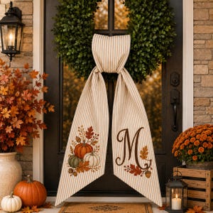 May include: A beige and white striped fabric door hanger featuring a green boxwood wreath at the top. One tail displays a printed cluster of pumpkins and autumn leaves, while the other tail features a large brown letter M with leaf accents.