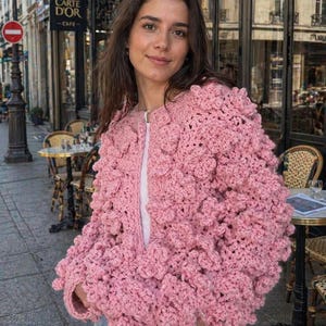 May include: A light pink, handmade crochet jacket with a textured, floral-like design. The jacket has a zipper closure and is worn over a white top. The jacket's unique texture and color make it a standout piece.