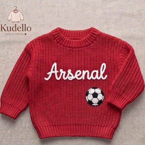 Pode incluir: Um suéter de malha vermelho brilhante de manga comprida com textura canelada, gola redonda e punhos canelados. A frente exibe o texto bordado em branco Arsenal em letra cursiva, posicionado acima de um emblema de bola de futebol.