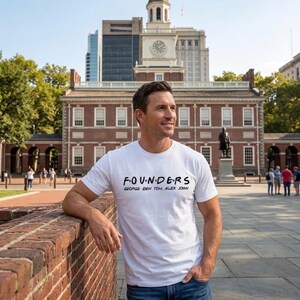 Op de afbeelding: Een wit t-shirt met het woord "FOUNDERS" in zwarte letters, met daaronder de namen George, Ben, Tom, Alex en John. Het shirt wordt gedragen door een persoon voor een bakstenen gebouw.