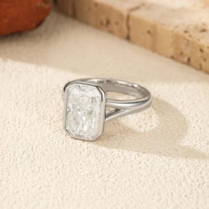 May include: A silver-toned ring featuring a large, rectangular, radiant-cut clear stone set in a bezel setting. The band is smooth and polished, with a split-shank design that connects to the central stone setting on both sides.