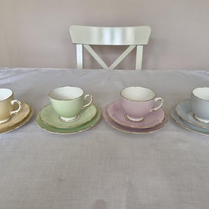 May include: A set of four porcelain teacups and saucers in pastel yellow, light green, soft pink, and pale blue. Each cup features a rounded bowl, a small handle, and a gold-trimmed rim, matching the circular saucer with a gold-trimmed edge.
