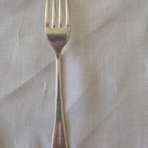 May include: A silver-toned metal dinner fork with four tines and a smooth, tapered handle. The handle features a small, vertical engraved mark near the base. The fork has a polished, reflective surface and a simple, clean design throughout its length.