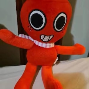 May include: A plush toy with a bright orange body, large circular eyes with black pupils, and a wide mouth showing silver teeth. It wears a light blue bow on its head and a pink and white checkered scarf around its neck, with red circular cheeks.