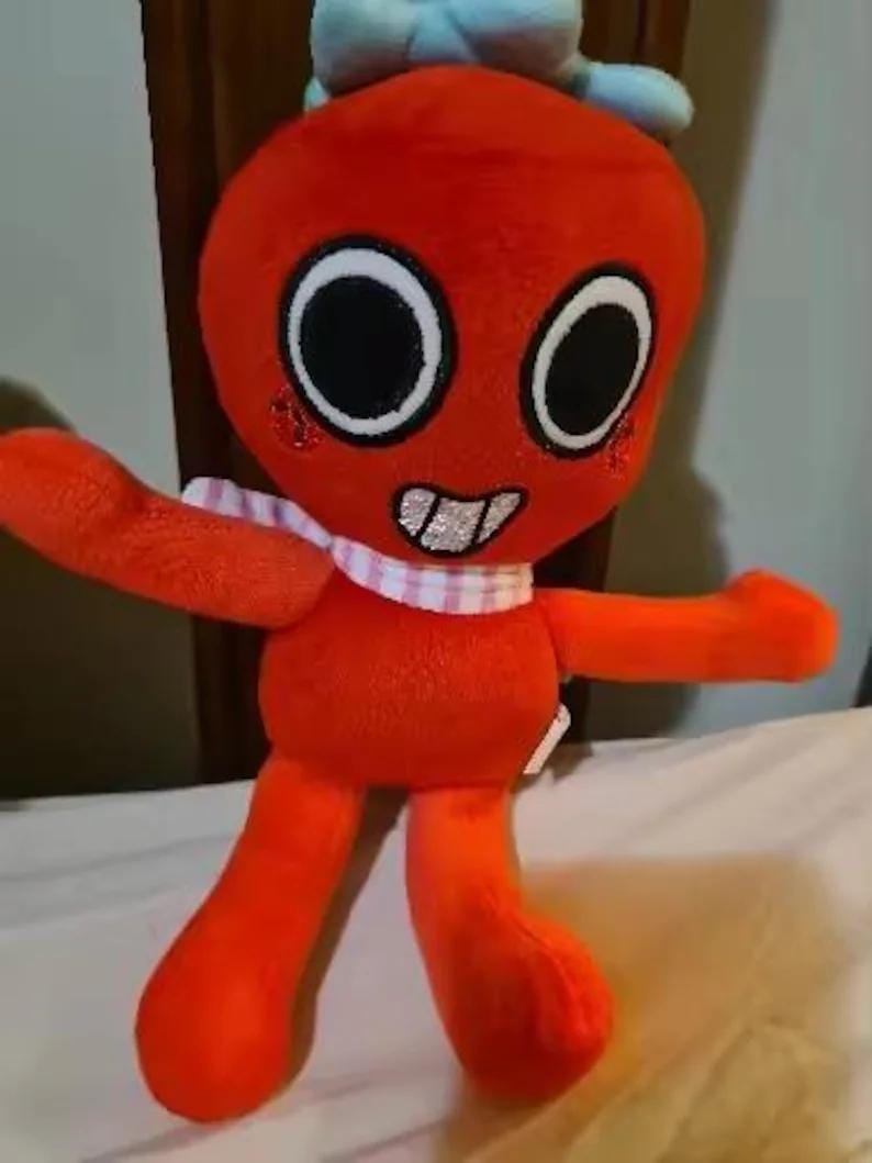 May include: A plush toy with a bright orange body, large circular eyes with black pupils, and a wide mouth showing silver teeth. It wears a light blue bow on its head and a pink and white checkered scarf around its neck, with red circular cheeks.