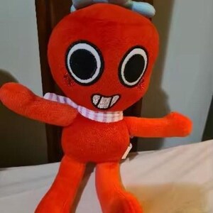 May include: A plush toy featuring a bright orange, round-headed character with large black eyes, a wide grin showing white teeth, and a light blue bow on top. The character wears a white and pink checkered scarf around its neck and has red cheeks.