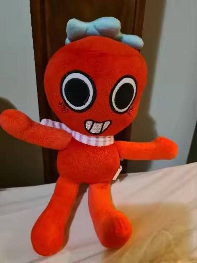May include: A plush toy featuring a bright orange, round-headed character with large black eyes, a wide grin showing white teeth, and a light blue bow on top. The character wears a white and pink checkered scarf around its neck and has red cheeks.
