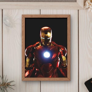 以下が含まれることがあります： 赤と金のアーマーを着たアイアンマンの額装プリント。胸には光る部分があります。「MOVIE」の文字がフレームの上に表示されています。画像はポップコーン、コーヒーカップ、リモコンに囲まれています。