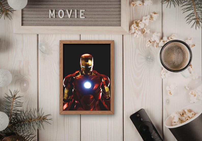 以下が含まれることがあります： 赤と金のアーマーを着たアイアンマンの額装プリント。胸には光る部分があります。「MOVIE」の文字がフレームの上に表示されています。画像はポップコーン、コーヒーカップ、リモコンに囲まれています。