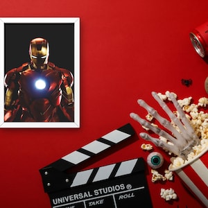 以下が含まれることがあります： 赤い背景に、アイアンマンの額縁入り画像と映画の小道具が並んでいます。 骸骨の手が付いたポップコーンバケツ、3Dメガネ、映画のクラッパーボードも存在します。 クモのイラストが描かれた赤い缶も表示されています。