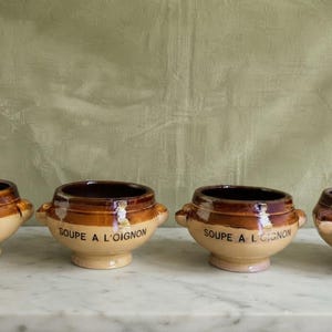 Peut inclure: Un ensemble de quatre bols en céramique avec deux petites anses chacun. Les bols présentent un design bicolore avec une base beige clair et un bord émaillé marron foncé. Le texte "SOUPE A L'OIGNON" est imprimé en noir sur chaque bol.