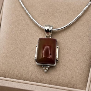 Op de afbeelding: Een zilveren ketting met een rechthoekige, diep roodbruine stenen hanger. De hanger is gezet in een gepolijst zilveren frame met kleine decoratieve metalen kraaltjes aan de onderkant. De ketting wordt gepresenteerd in een beige gevoerd juwelendoosje.