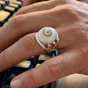 Peut inclure: Une bague en argent avec une grosse pierre ronde de couleur crème avec un motif en spirale. La bague est portée au doigt d'une personne, avec un clavier noir au premier plan. La bague a un design ajouré sur les côtés.