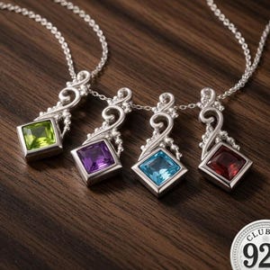 Può includere: Quattro collane d'argento con pendenti quadrati con pietre preziose verdi, viola, blu e rosse. Ogni pendente è incastonato in una montatura d'argento decorativa e pende da una delicata catena. L'immagine è su una superficie di legno scuro.