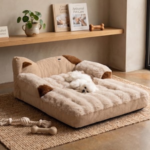 Peut inclure: Un lit pour chien beige avec une surface de couchage en fausse fourrure moelleuse. Le lit a un extérieur en velours côtelé et des empiècements bruns. Un petit chien blanc se repose sur le lit. Un jouet en corde et un jouet en forme d'os sont au premier plan.