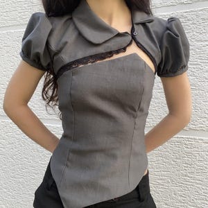 May include: A gray top with a unique design. It features short, puffed sleeves, a collared neckline, and a cutout detail at the chest, trimmed with black lace. The top is paired with black pants.