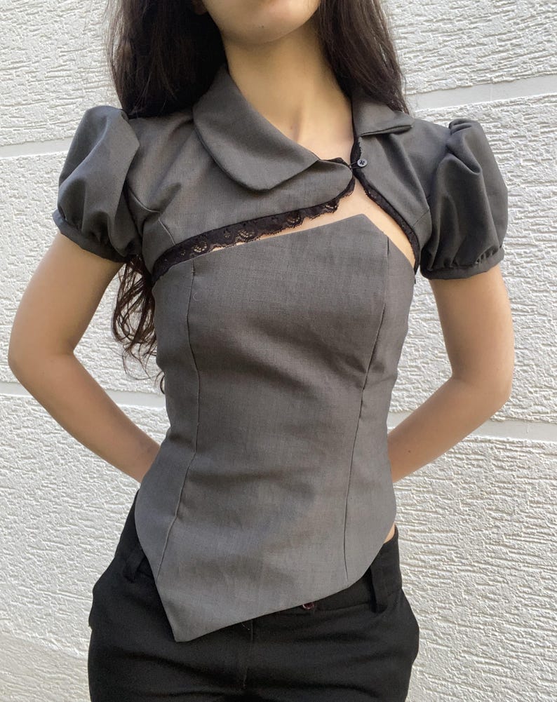 May include: A gray top with a unique design. It features short, puffed sleeves, a collared neckline, and a cutout detail at the chest, trimmed with black lace. The top is paired with black pants.