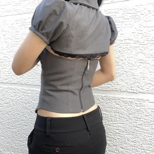 May include: Gray top with short, puffed sleeves, a collar, and a black lace trim. The top has a zipper down the back. The model is also wearing black pants.