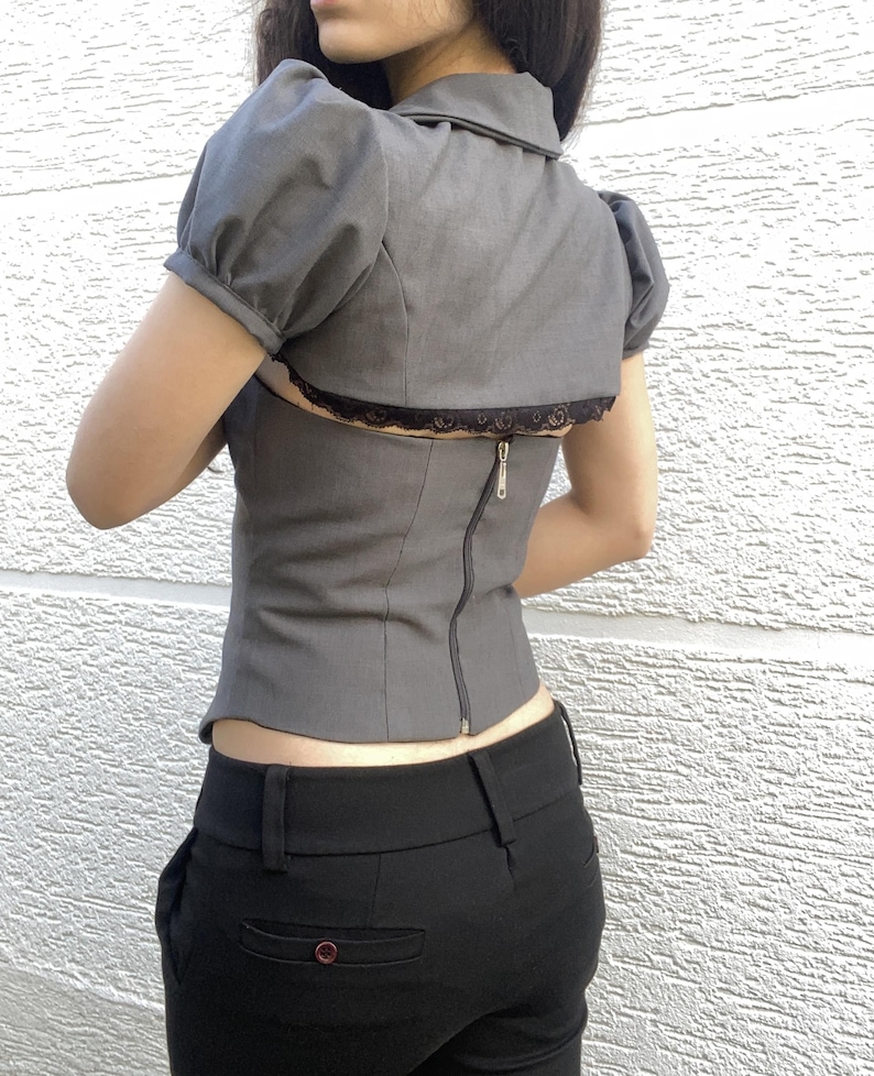 May include: Gray top with short, puffed sleeves, a collar, and a black lace trim. The top has a zipper down the back. The model is also wearing black pants.