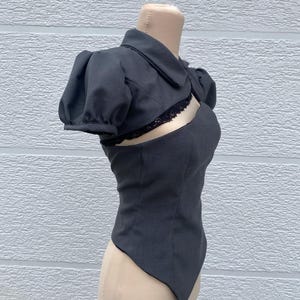 May include: A dark gray, two-piece top with a unique design. The top features a collared, short-sleeved bolero jacket and a fitted bodice with a cutout detail. Black lace trim accents the cutout. The garment is displayed on a mannequin.