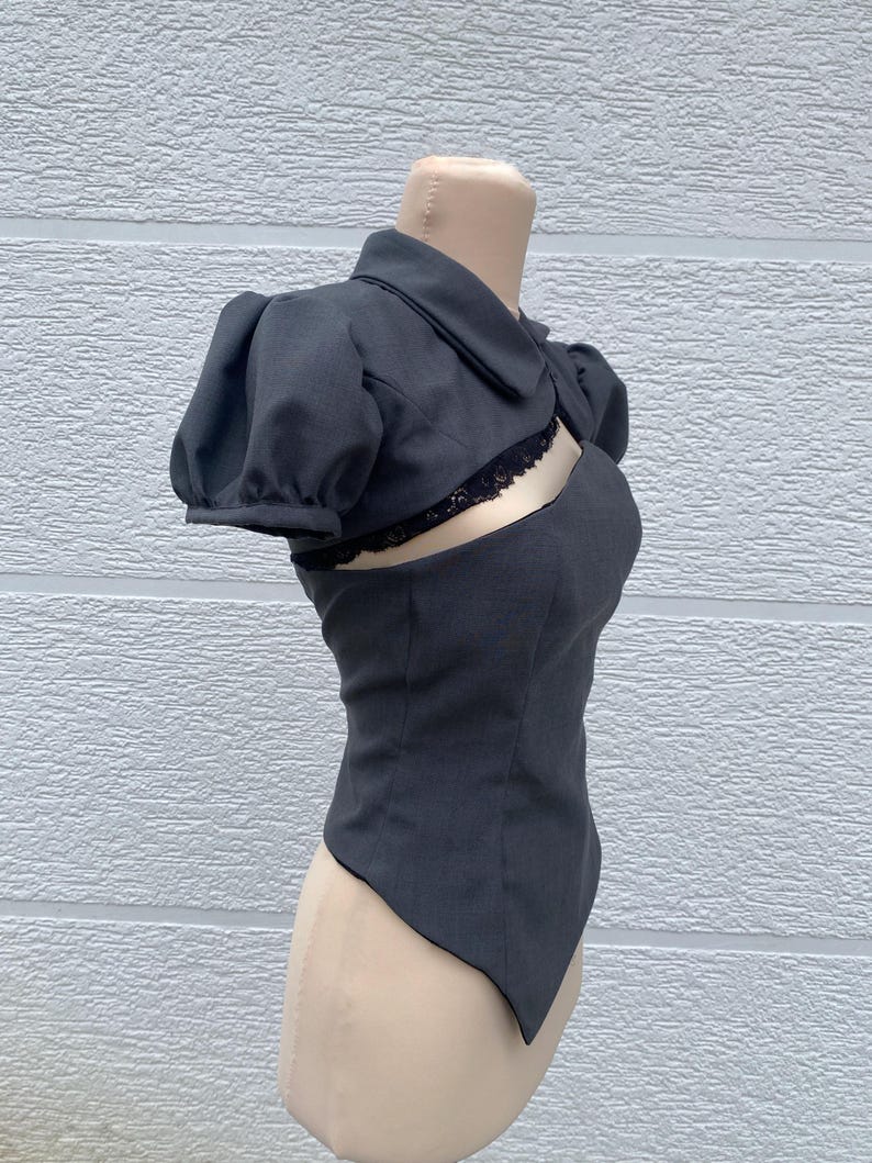 May include: A dark gray, two-piece top with a unique design. The top features a collared, short-sleeved bolero jacket and a fitted bodice with a cutout detail. Black lace trim accents the cutout. The garment is displayed on a mannequin.