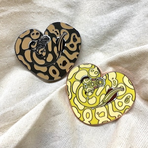 Peut inclure: Deux épingles en émail en forme de cœur représentant un serpent. La première épingle est noire et marron avec un fond marron clair. La deuxième épingle est jaune et blanche avec un fond jaune clair.