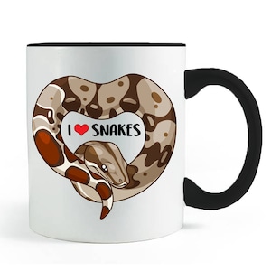 May include: White ceramic coffee mug with a black rim and handle. The mug features a cartoon image of a brown and tan snake curled into a heart shape with the text "I love snakes" in red.